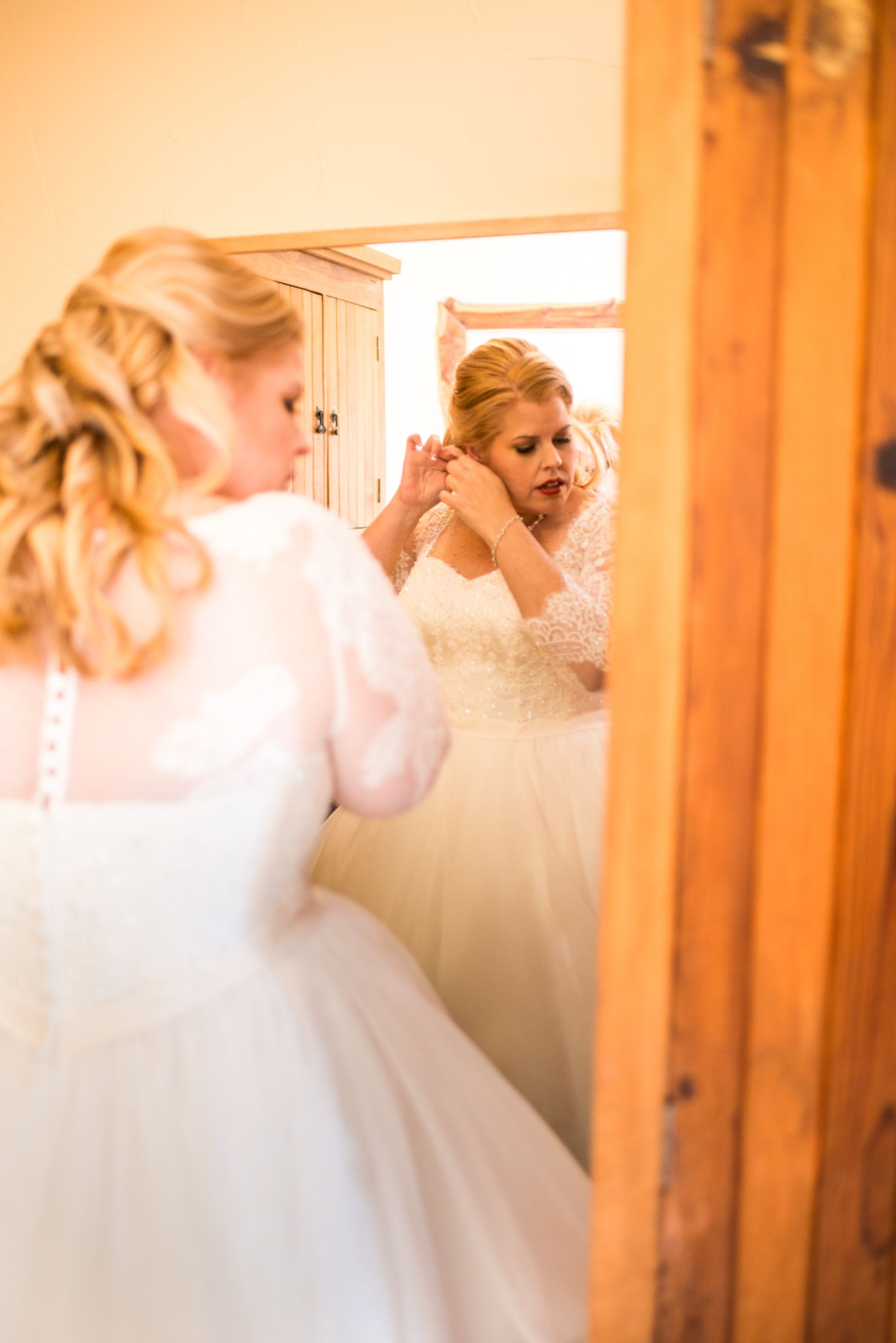 Bride in white dress putting on earrings while looking in mirror.