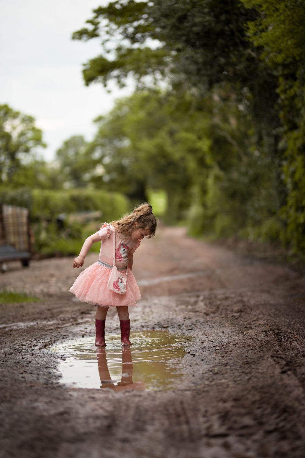 A child in a pink tutu splashes in puddles on a dirt path.