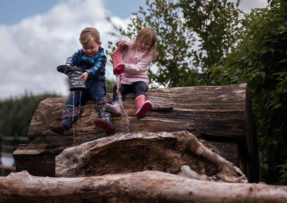 Children play on a large fallen log in a natural outdoor setting.