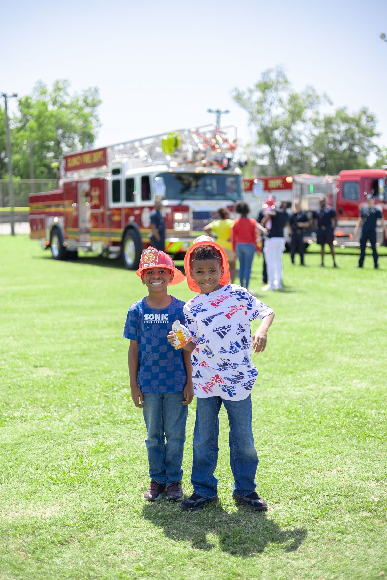 Two young boys standing together on grass with a fire truck visible in the background.