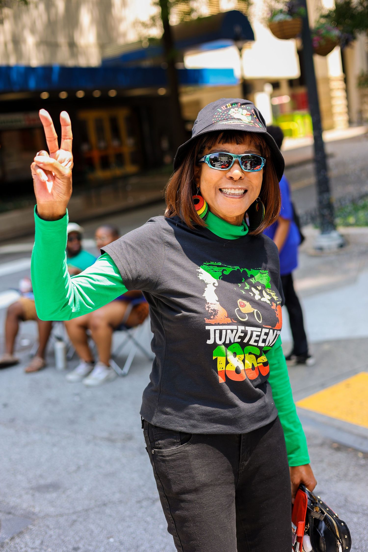 A person in a gray hat and green sleeves making a peace sign while standing on a city street.