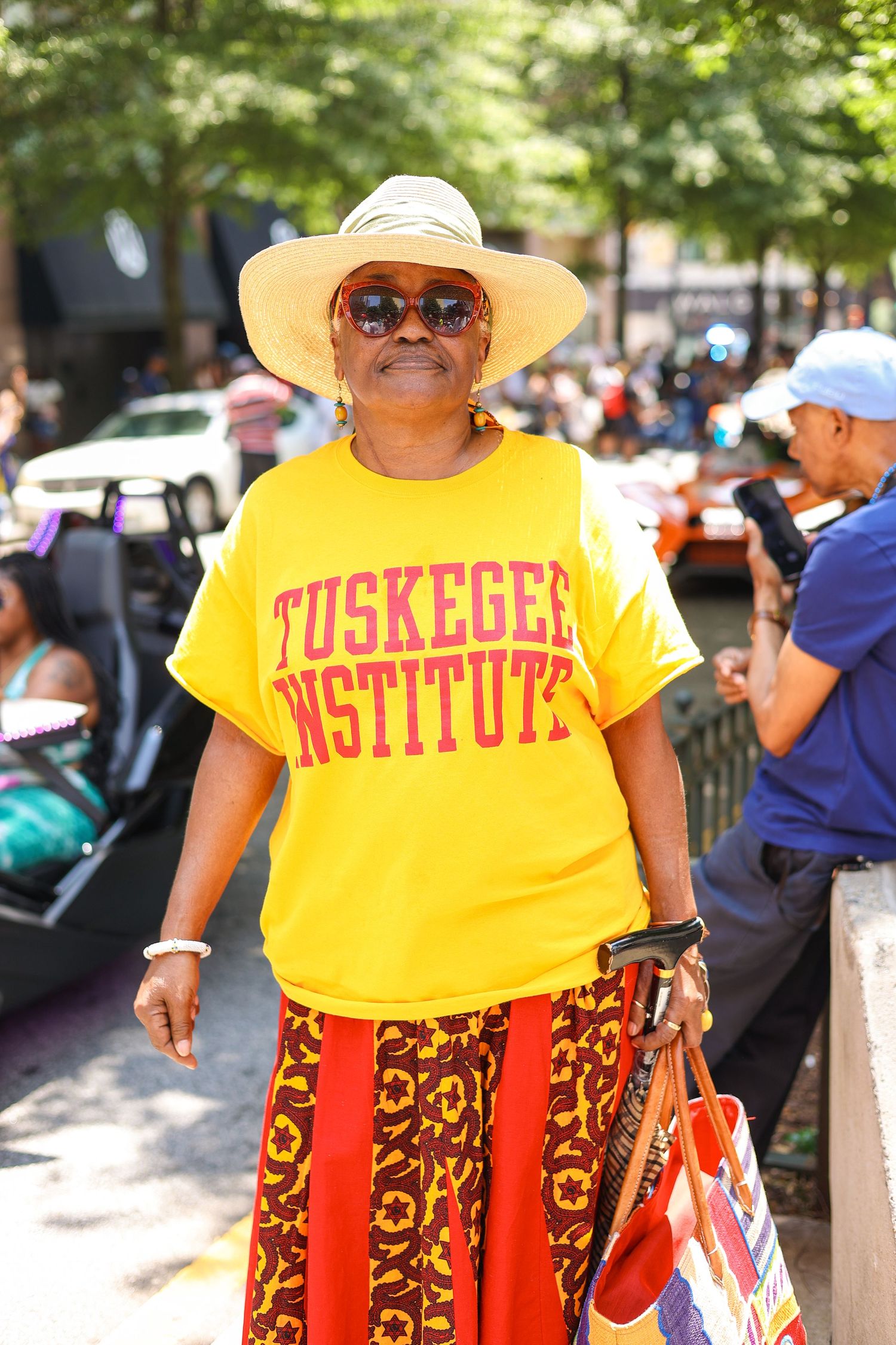A smiling person in a yellow Tuskegee Institute t-shirt, straw hat and patterned skirt enjoying an outdoor event.