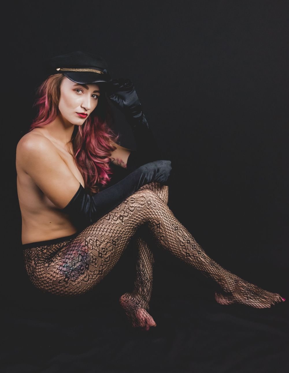 Woman with red hair wearing black cap, lace fishnet tights and gloves posing on dark studio background.
