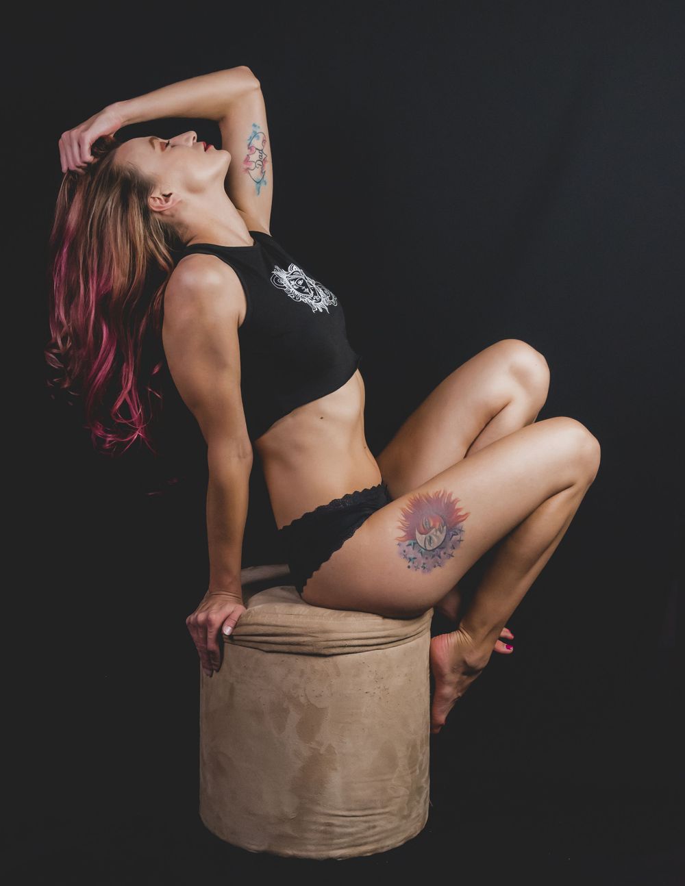 Red-haired woman with floral thigh tattoo in black crop top posing on stool against dark studio background.