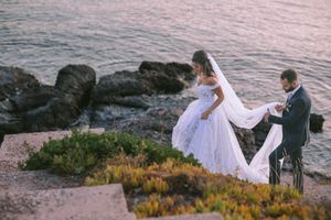 A couple in wedding attire standing by rocky coastal cliffs at sunset while the veil flows in the ocean breeze.