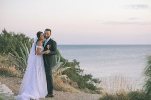 A couple in wedding attire embrace on a coastal cliff overlooking the ocean at sunset by GeorgeNafpliotis.com
