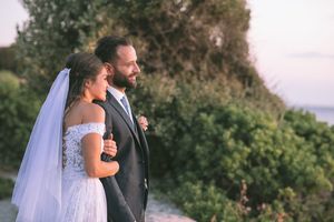 A couple in wedding attire embrace on a coastal cliff overlooking the ocean at sunset by GeorgeNafpliotis.com