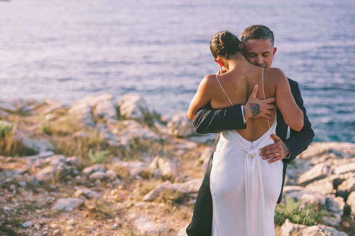 A couple in formal wedding on a rocky coastal cliff overlooking the blue ocean at sunset in Kefalonia captured By Georgenafpliotis.com
