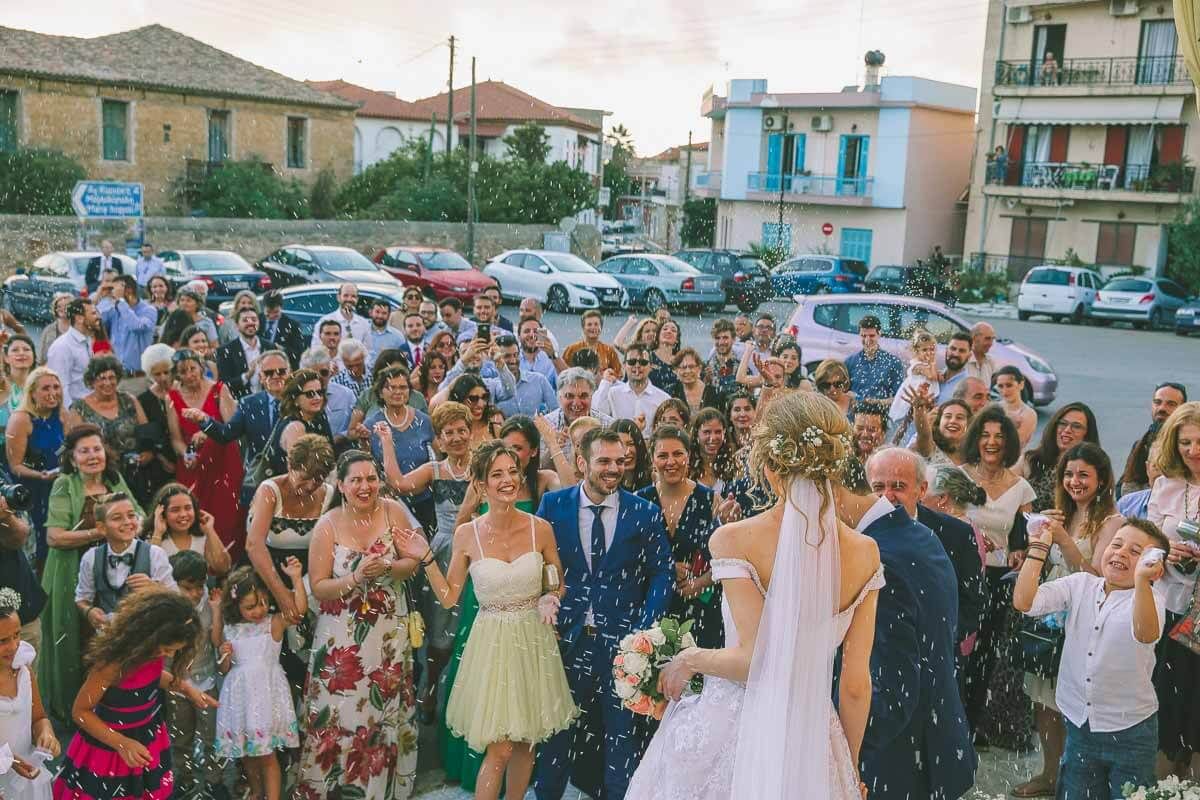 Crowd of wedding guests celebrating outdoors in a parking lot while throwing confetti on a sunny day in Greece.www.georgenafpliotis.com