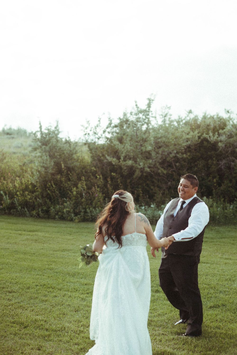 A couple dances together on a grassy lawn with shrubs in the background during a wedding celebration.