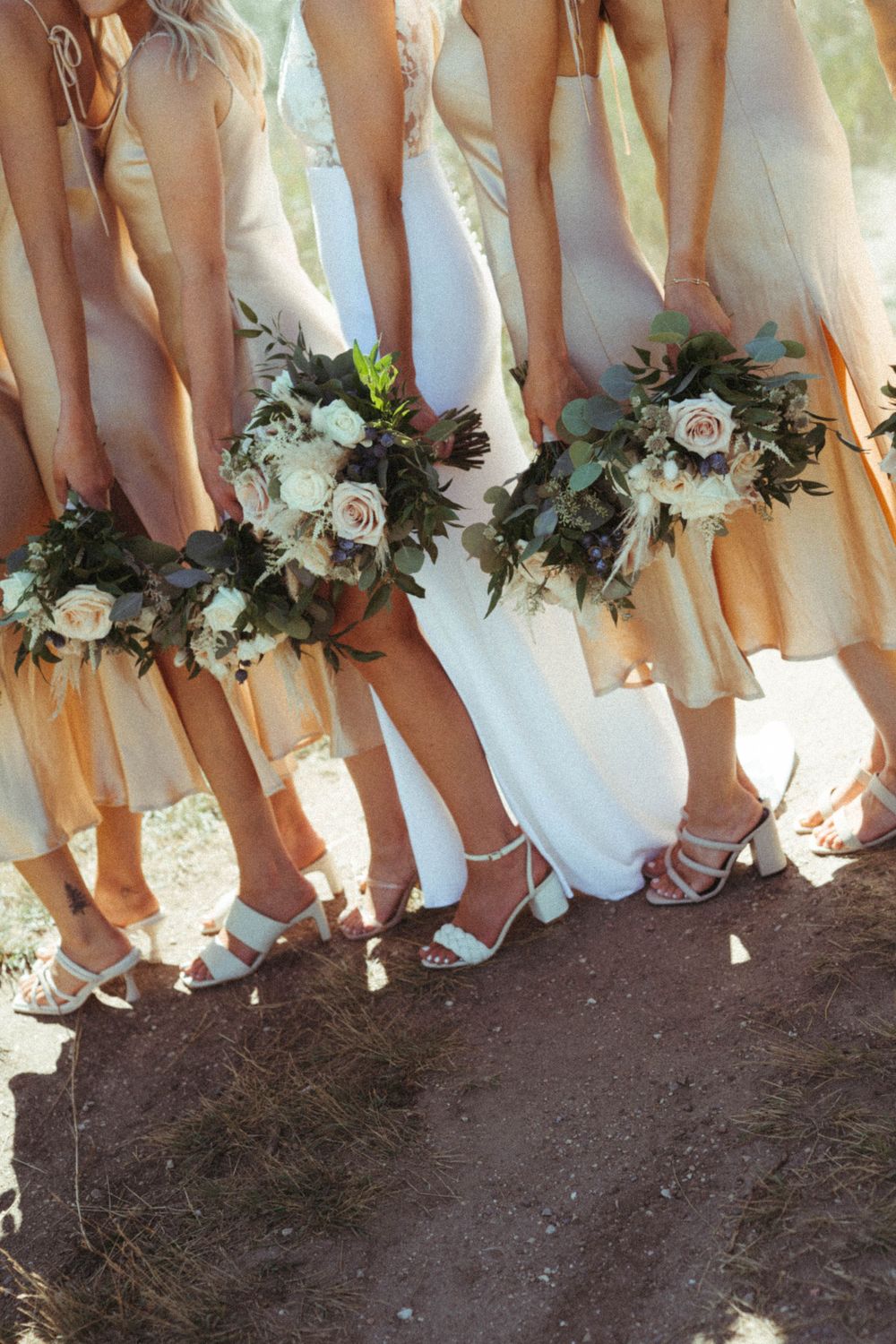 Bridesmaids in peachy dresses holding white floral bouquets show off their matching shoes in a candid wedding photo.