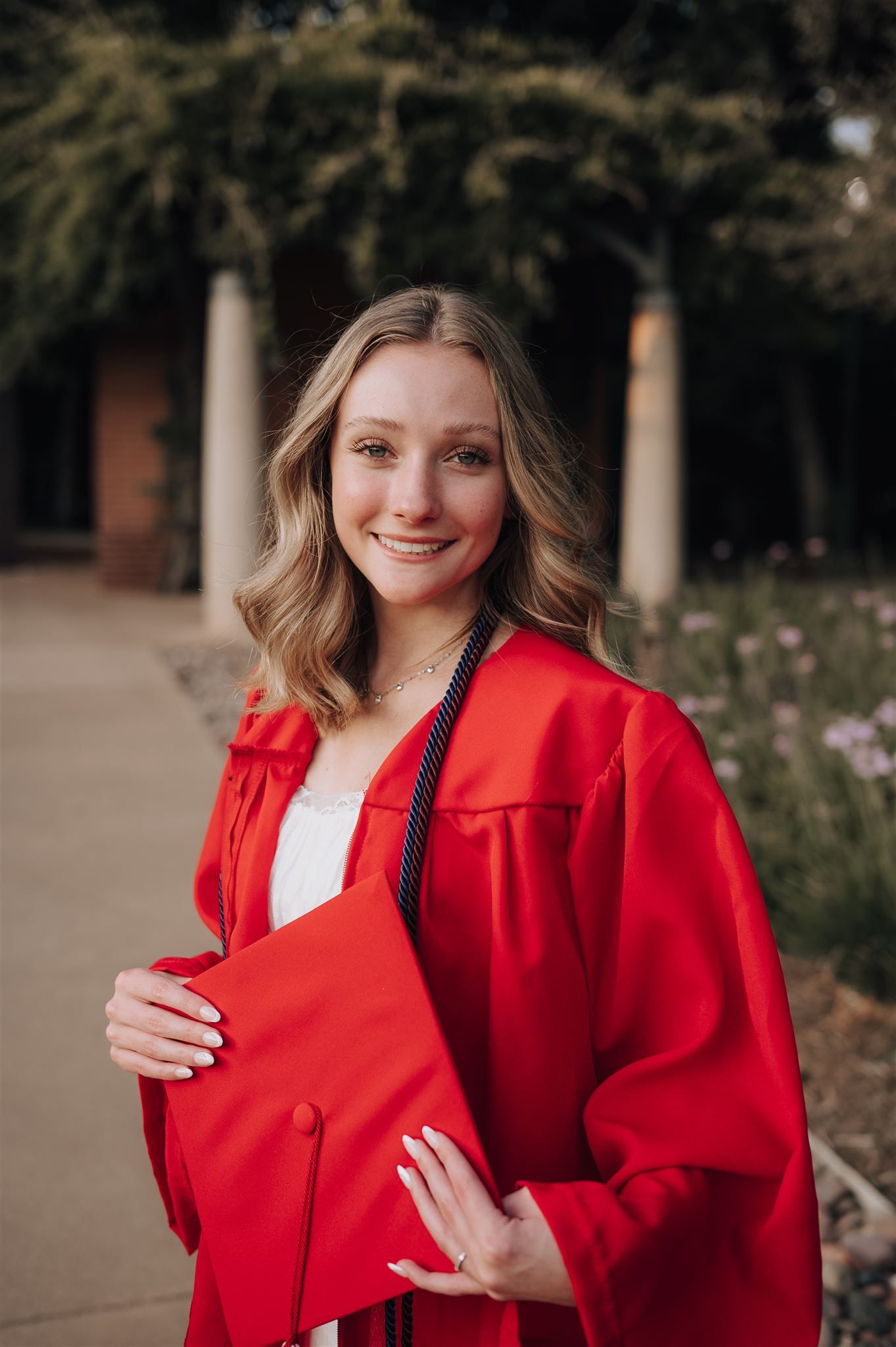 A graduate posing in a bright red graduation gown on a campus walkway.