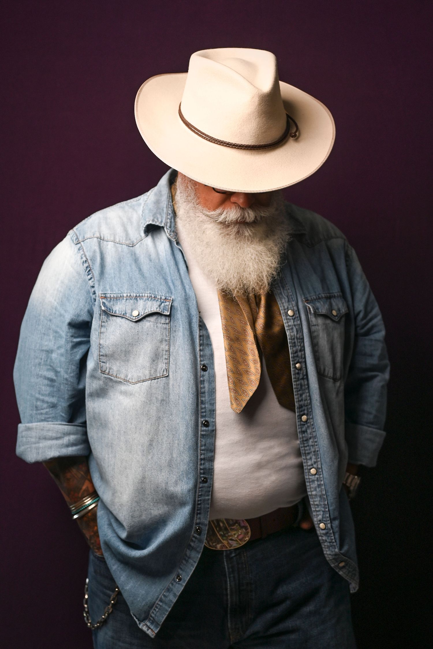 Portrait of person in denim jacket and cowboy hat with white beard against dark background.