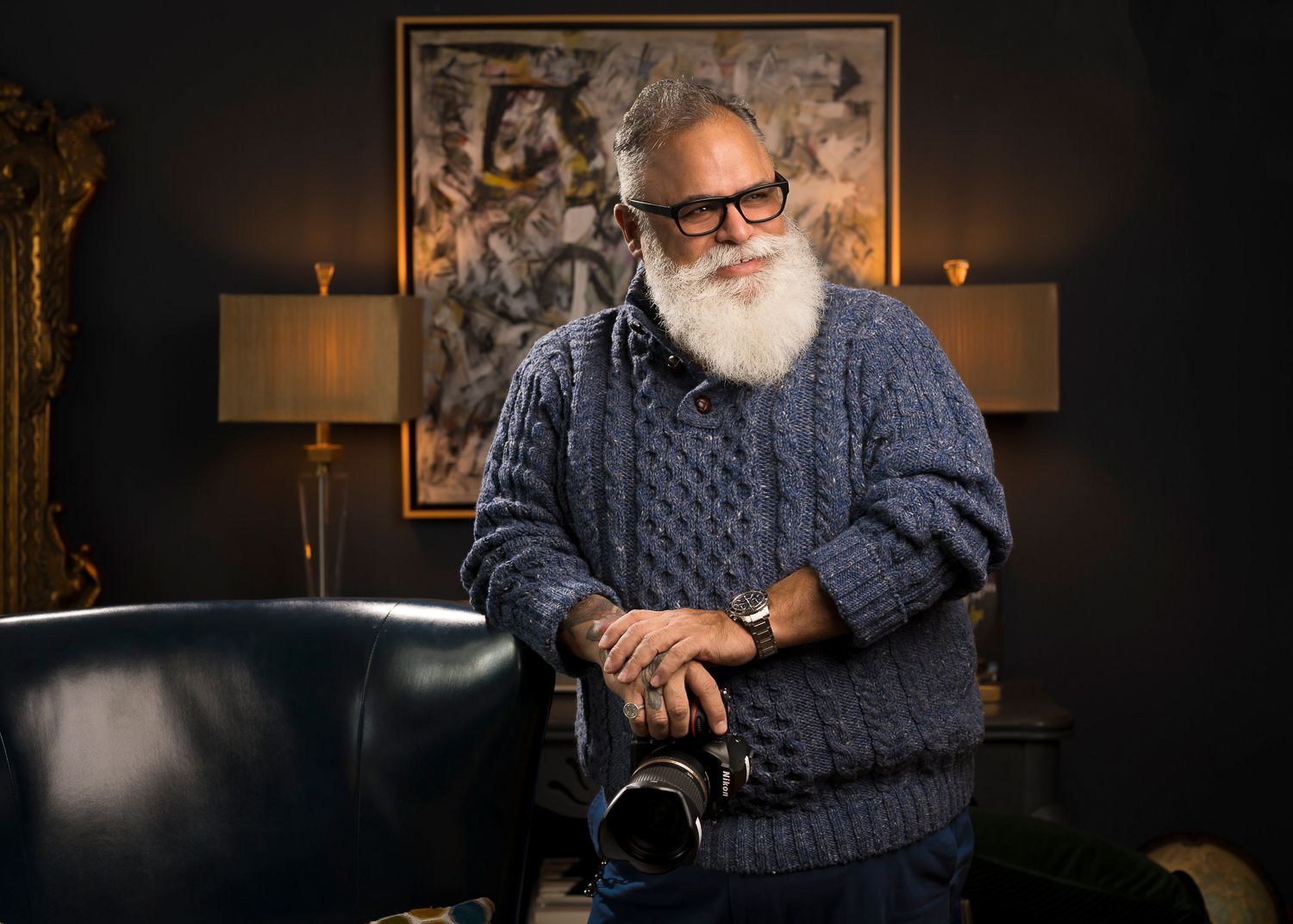 Portrait of person with white beard in cable knit sweater holding camera in elegant room setting.
