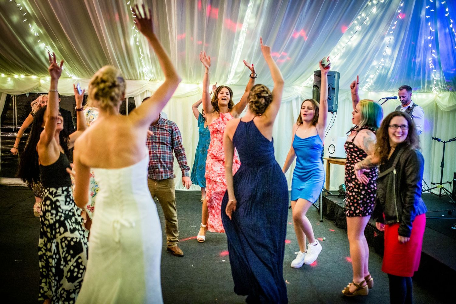 Wedding guests dancing energetically under colorful lighting in a reception tent.