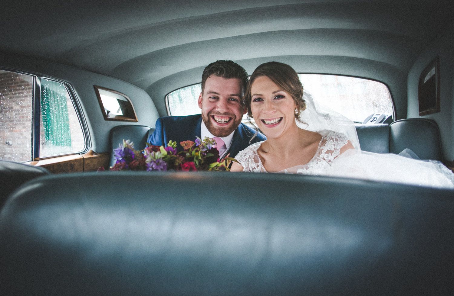 A wedding couple shares a joyful moment in the backseat of a classic car.