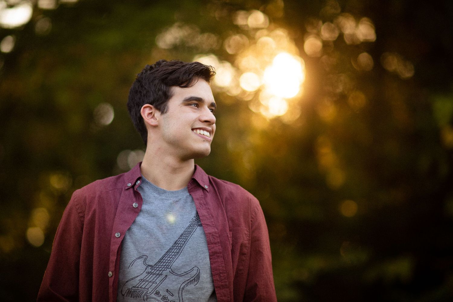Portrait of person in burgundy shirt against blurred sunset and autumn foliage.