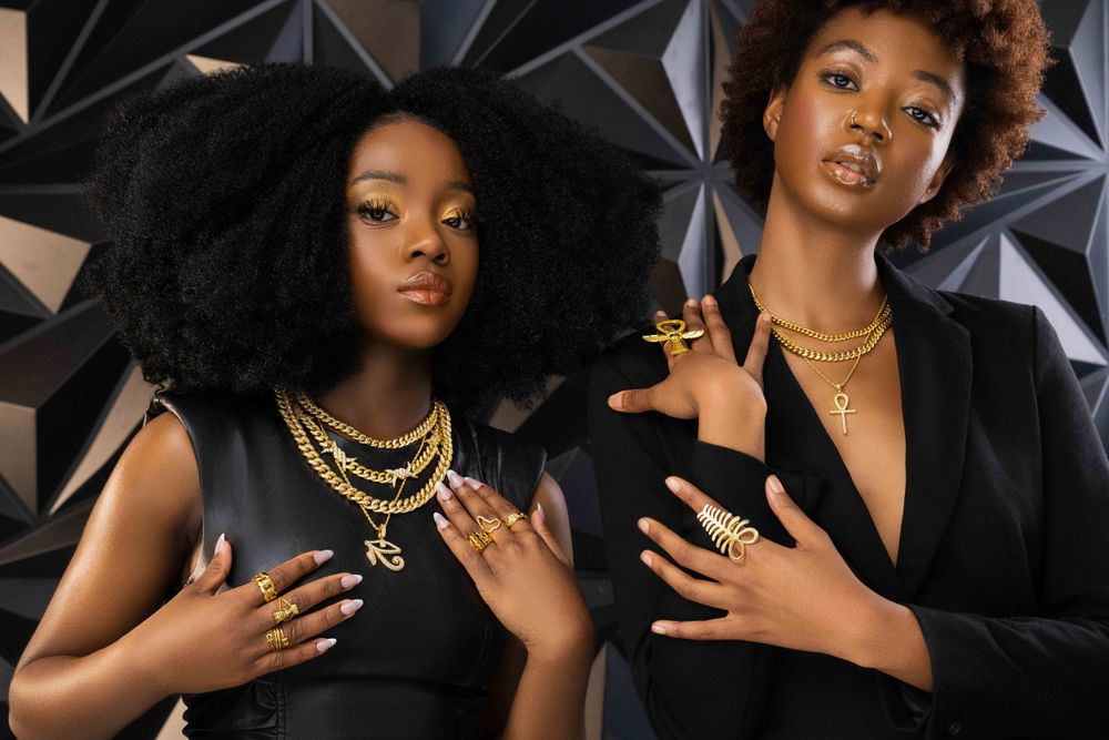 Two models in black outfits show off gold jewelry and professional manicures against geometric background.