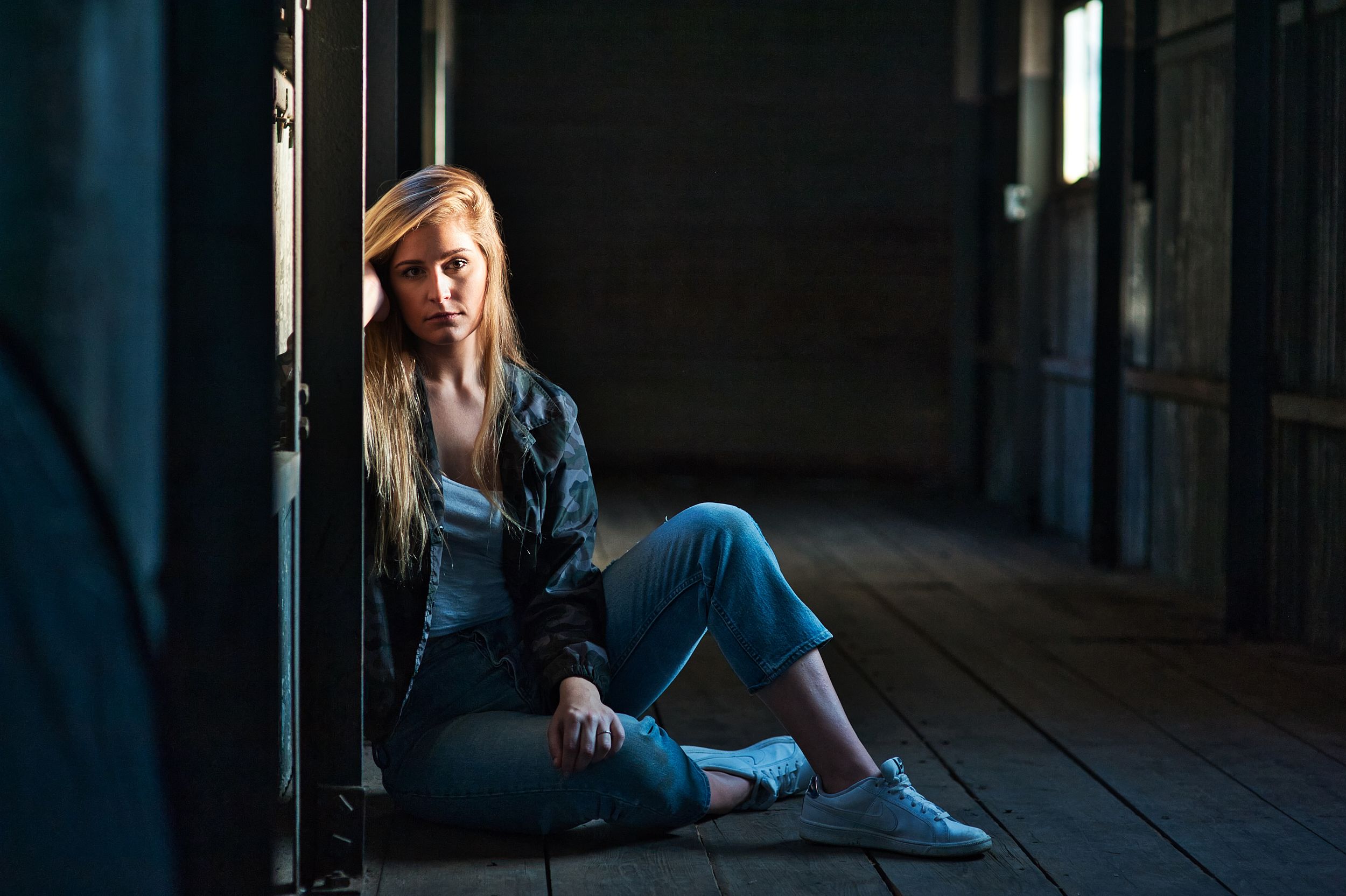 A blonde woman in jeans and jacket sits on a wooden floor in a dimly lit hallway.