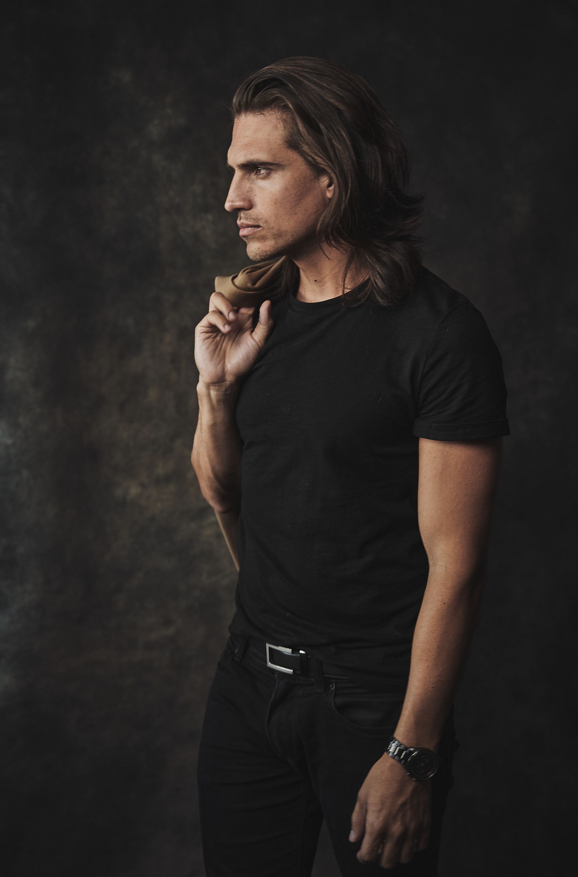 A fit man with long blonde hair in a black t-shirt poses against a dark background.