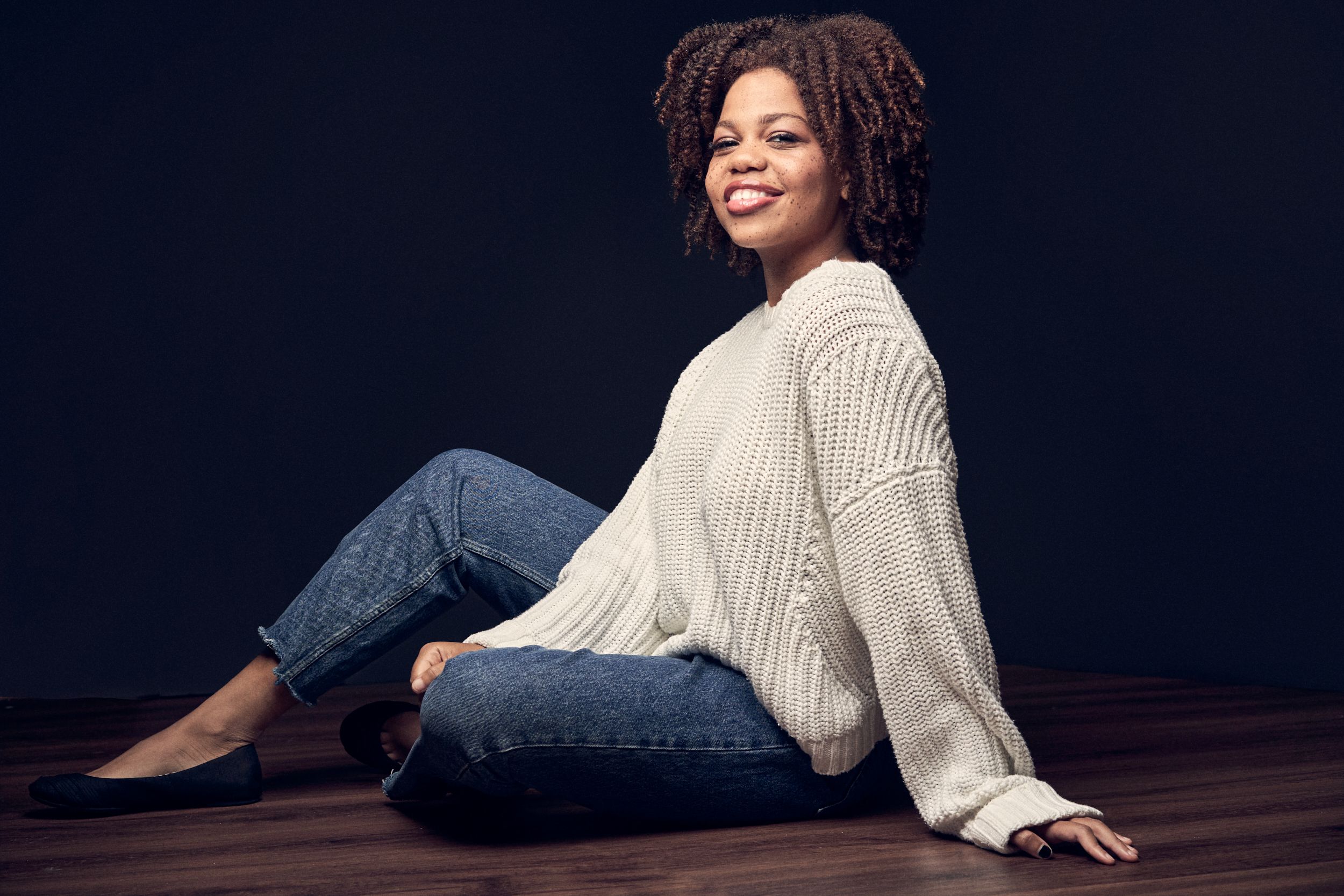 A smiling woman in a cream sweater and jeans sits relaxed on a wooden floor.