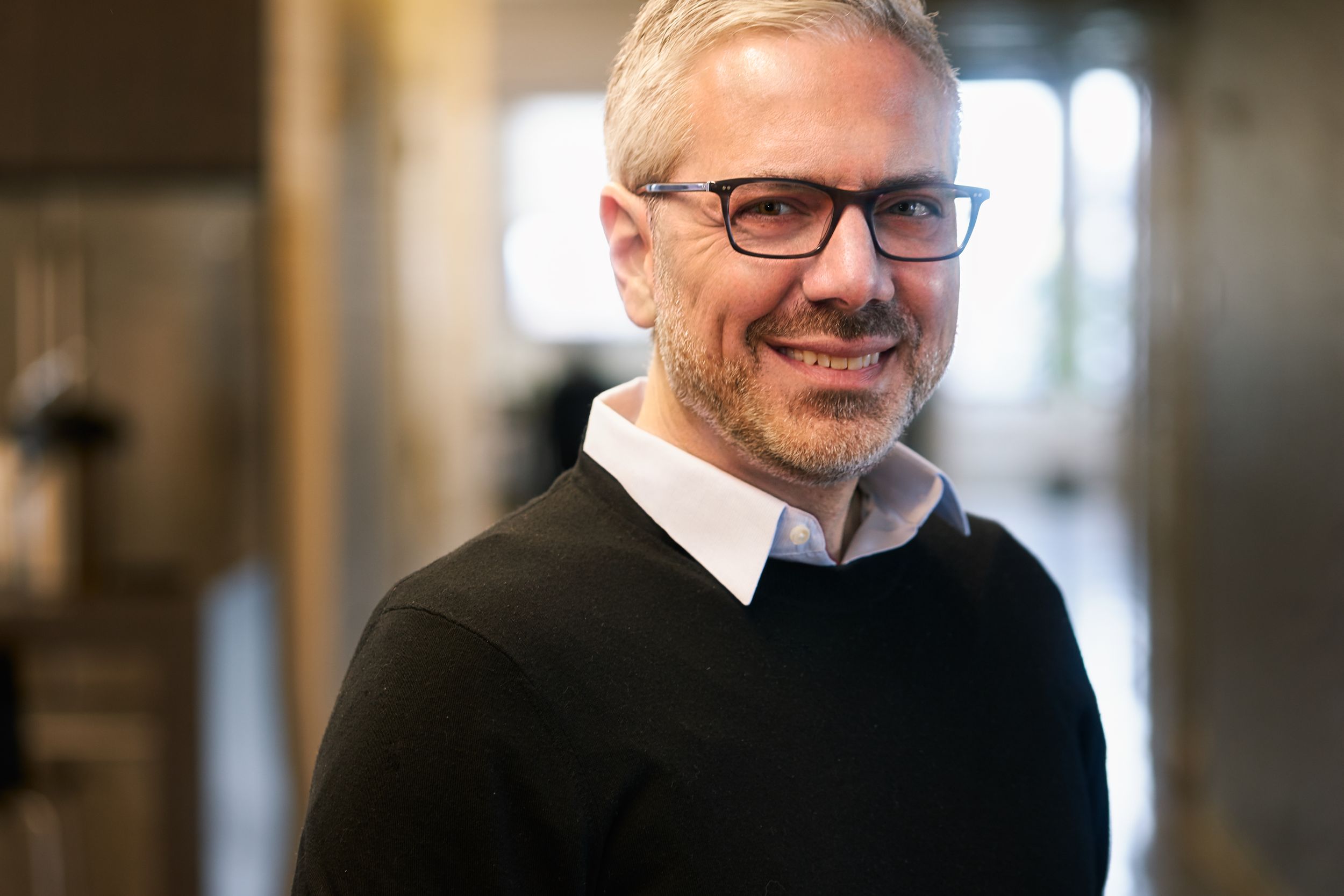A smiling middle-aged man with gray hair and glasses wearing a black sweater indoors.