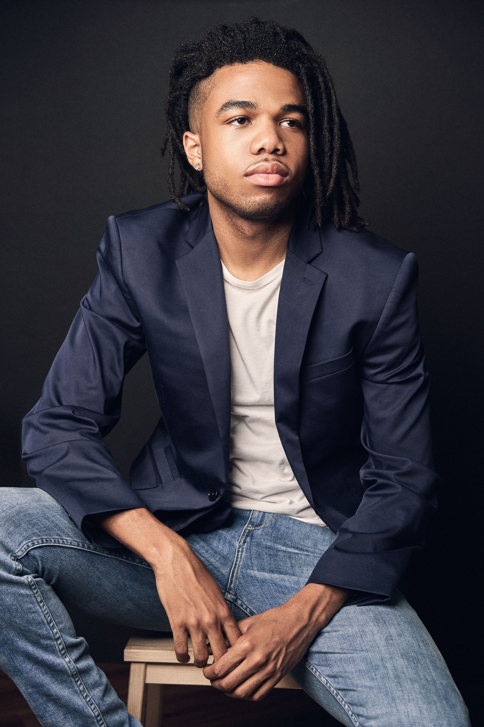 A young man with dreadlocks in a navy blazer and jeans poses confidently on a stool.
