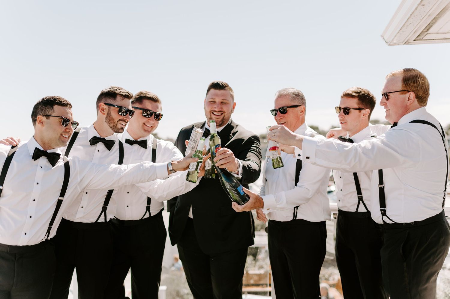 Groomsmen in white shirts and suspenders celebrate with the groom in black tuxedo outdoors.