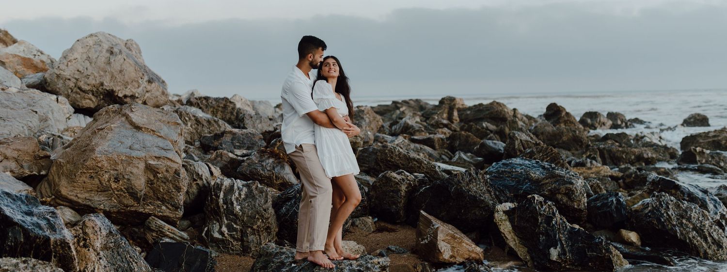 An engaged couple looks into each other's eyes as they stand together on a rocky beach overlooking the ocean.