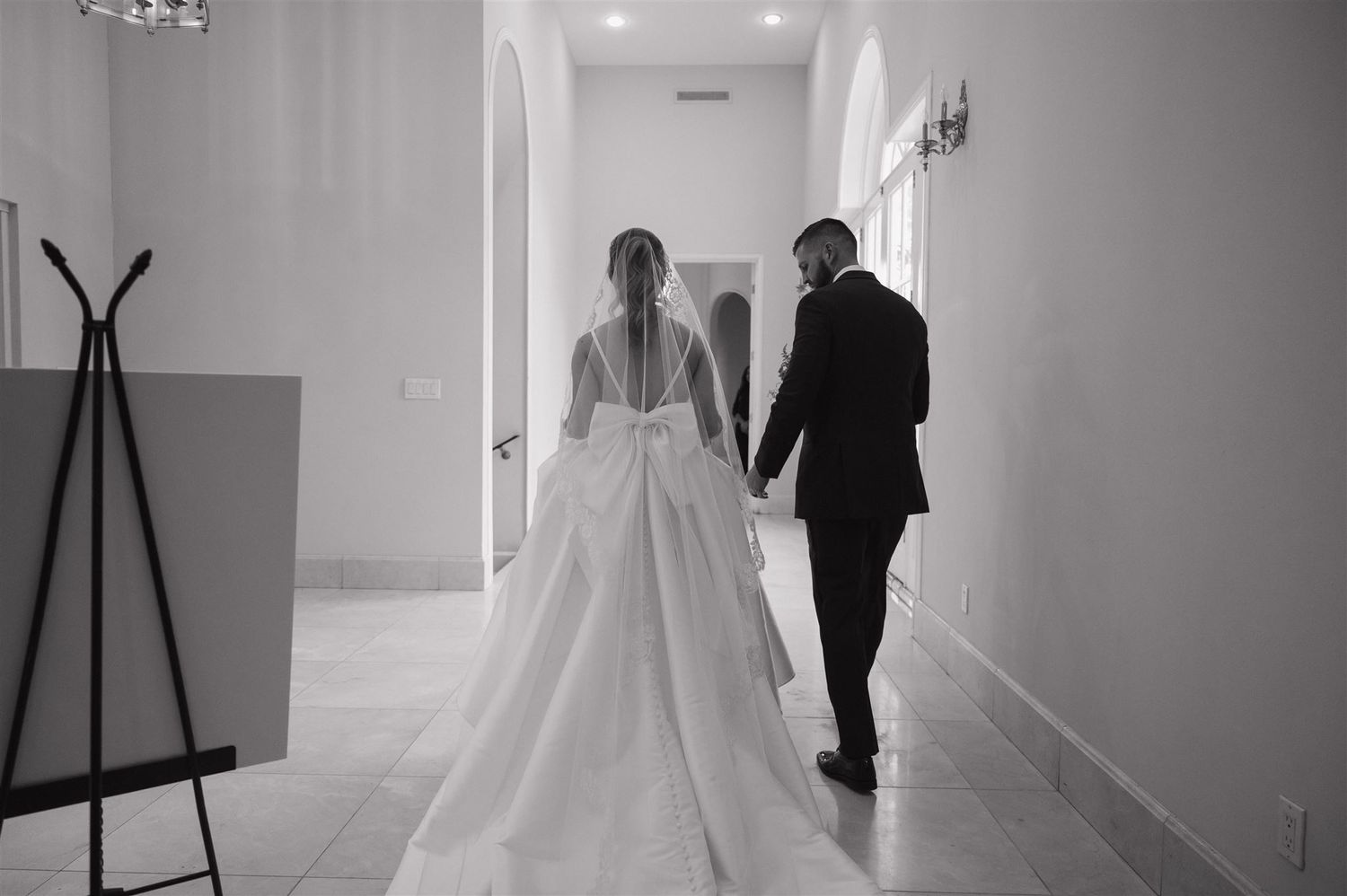 A romantic black and white photo of a couple walking hand in hand down a hallway at their wedding.