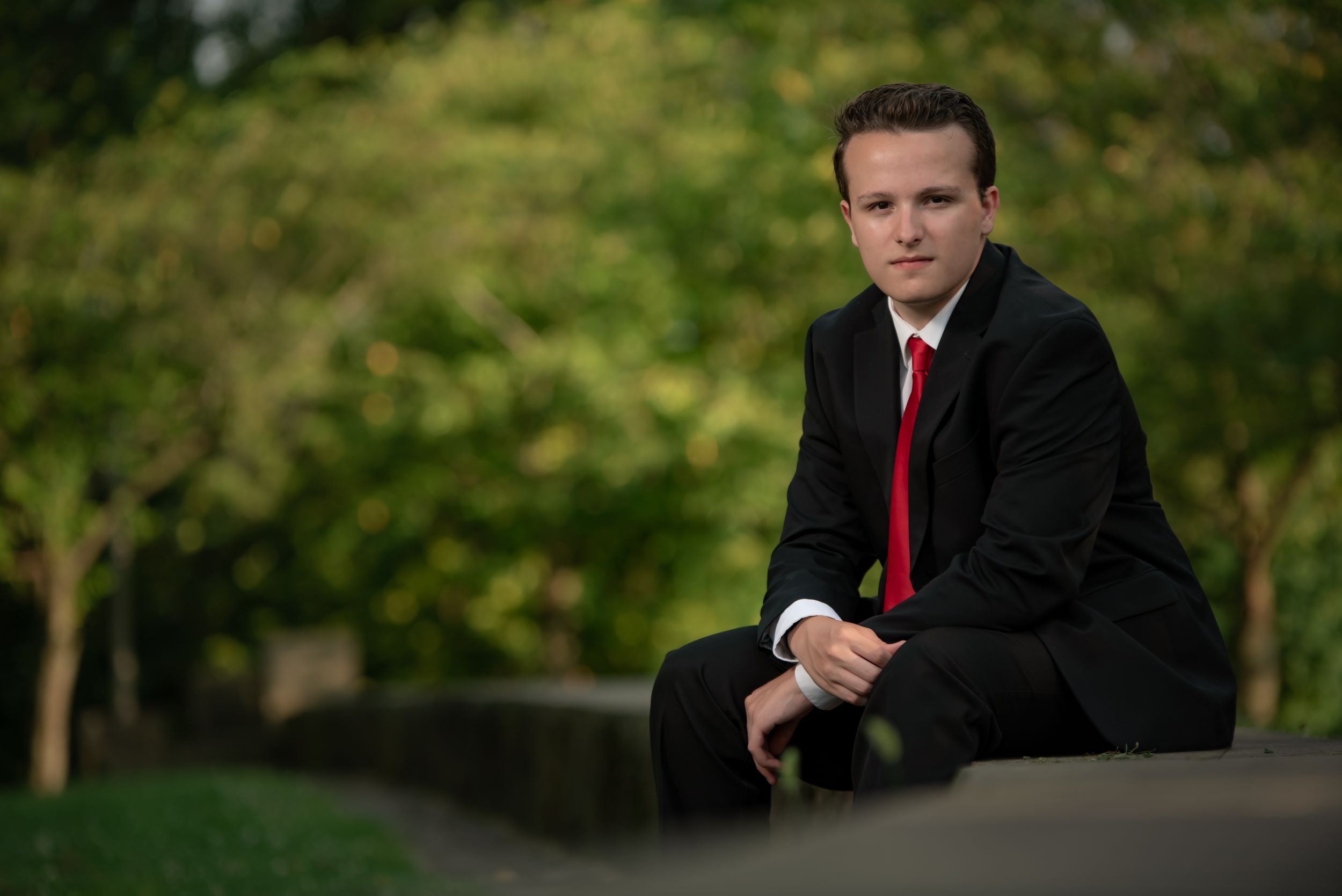 A formal portrait of someone in a suit and red tie seated outdoors.