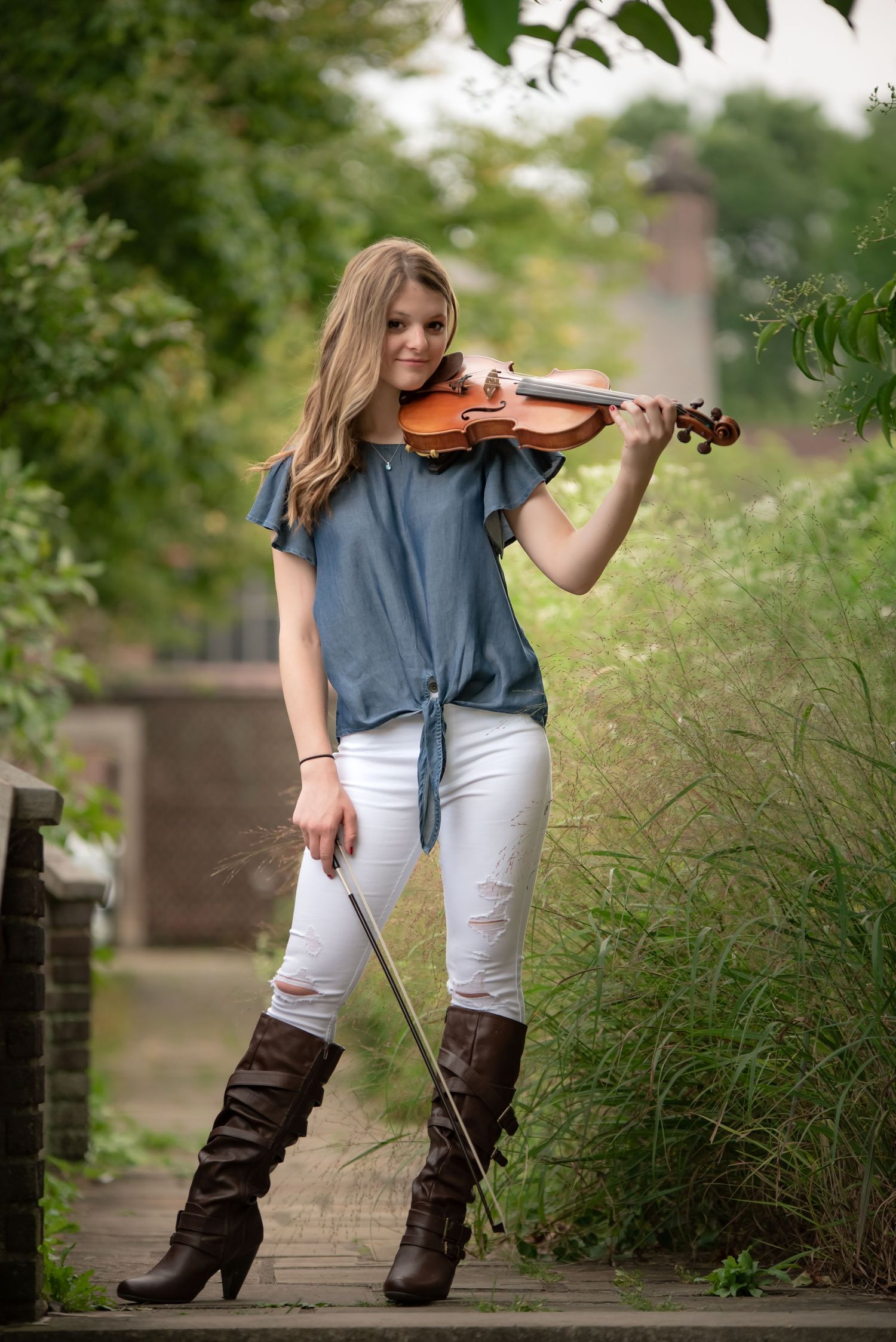 A violinist in casual attire and brown boots performs outdoors on a garden path.