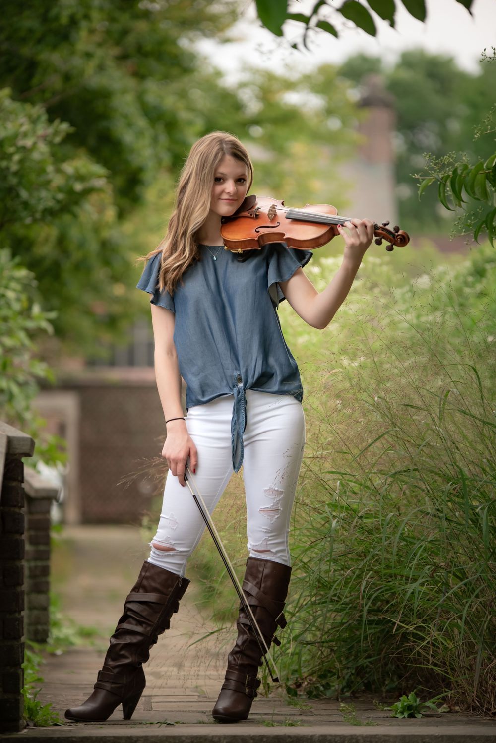 A violinist in casual attire and brown boots performs outdoors on a garden path.