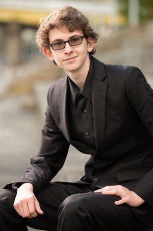 Professional portrait of a young person in glasses and black suit seated outdoors.