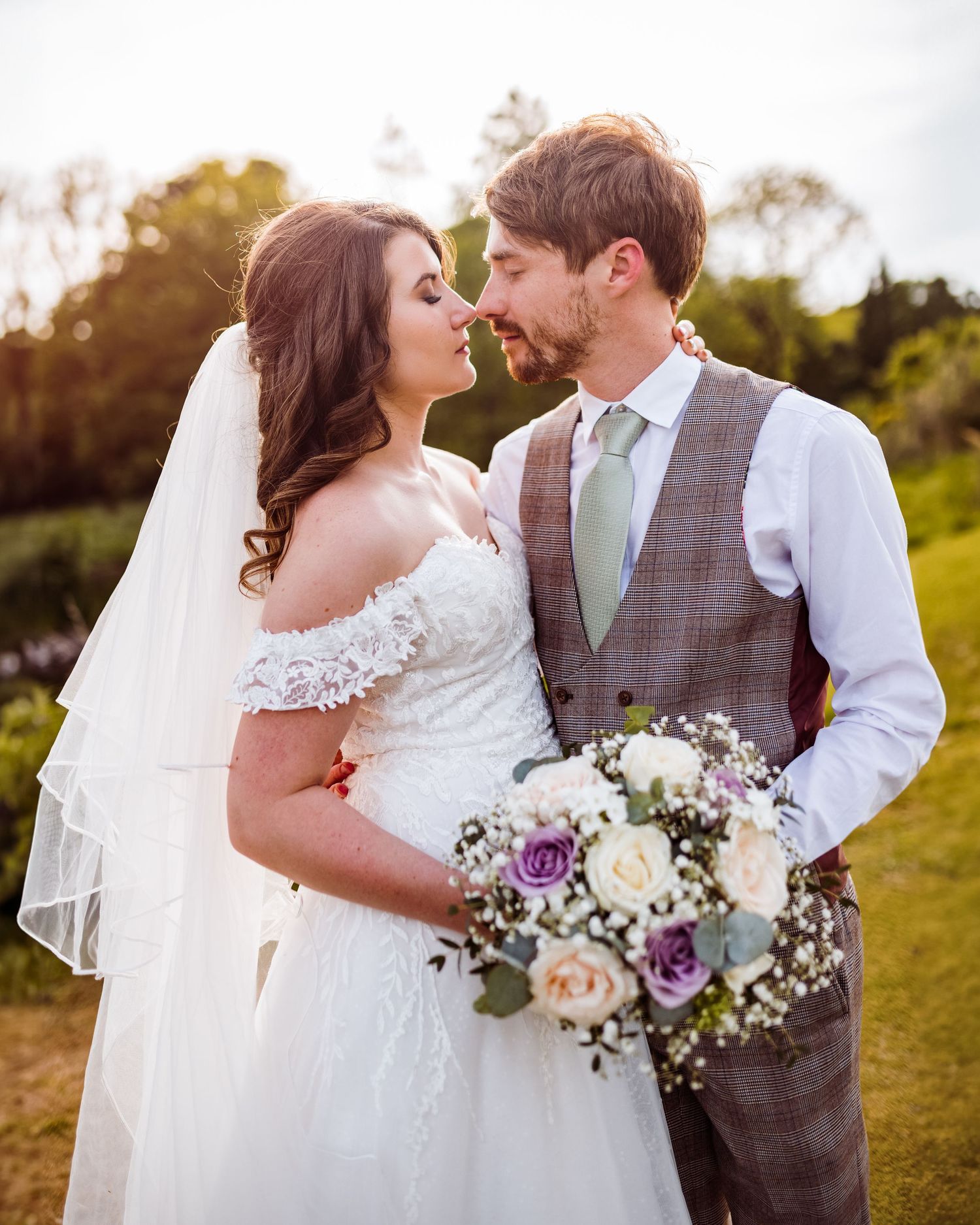Newlywed couple holding a bouquet of purple and white flowers in a scenic outdoor setting.