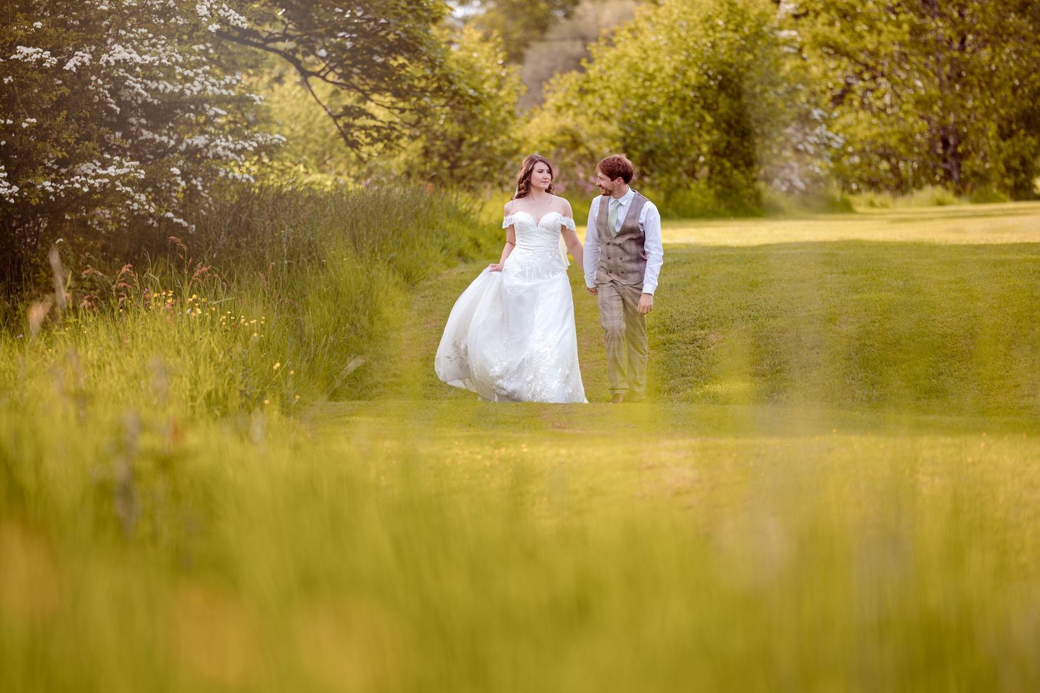 Newlyweds walk together through sunlit meadow during golden hour.
