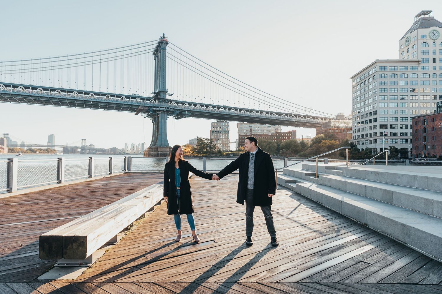 Couple holding hands on a wooden boardwalk with the Manhattan Bridge in the background.