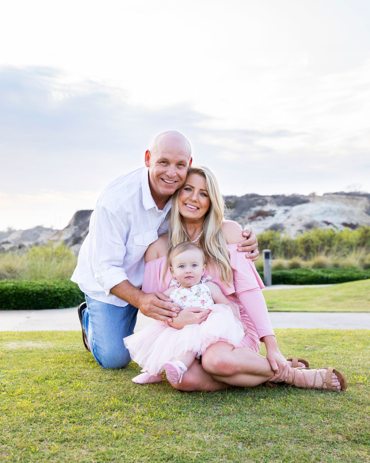 birthday photoshoot with Happy family of three posing on grass outdoors, parents smiling with baby girl in pink dress.