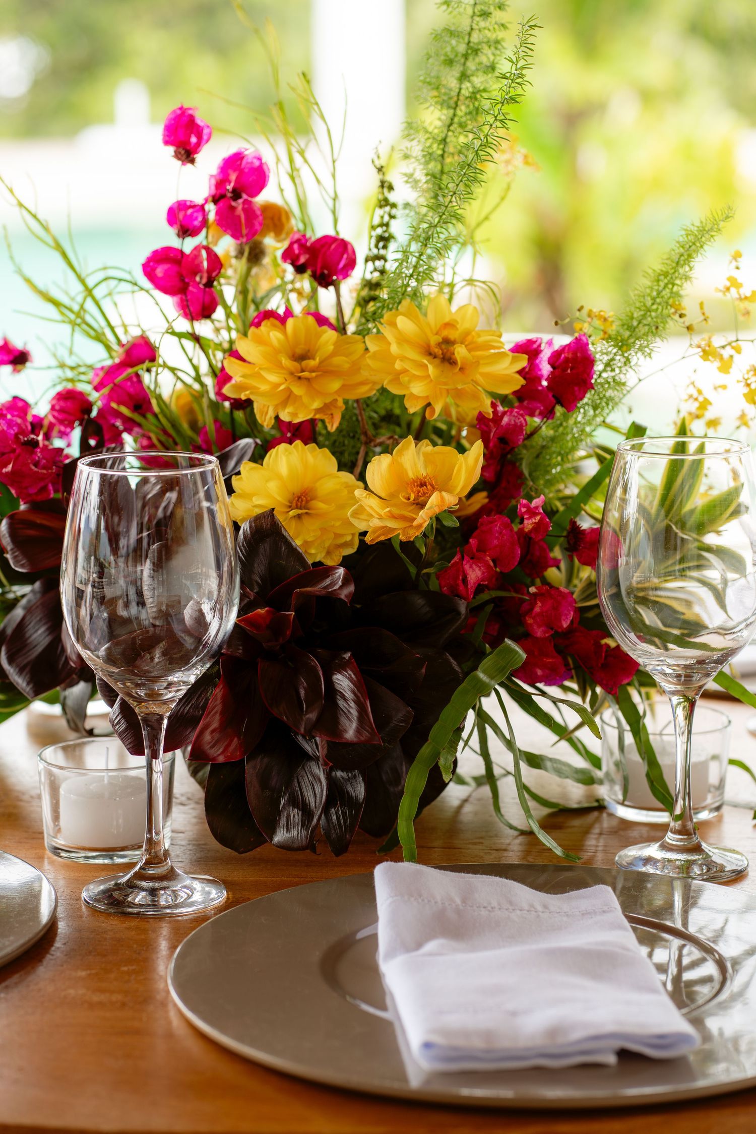 Colorful wedding centerpiece featuring yellow and pink flowers with wine glasses on a formal place setting.