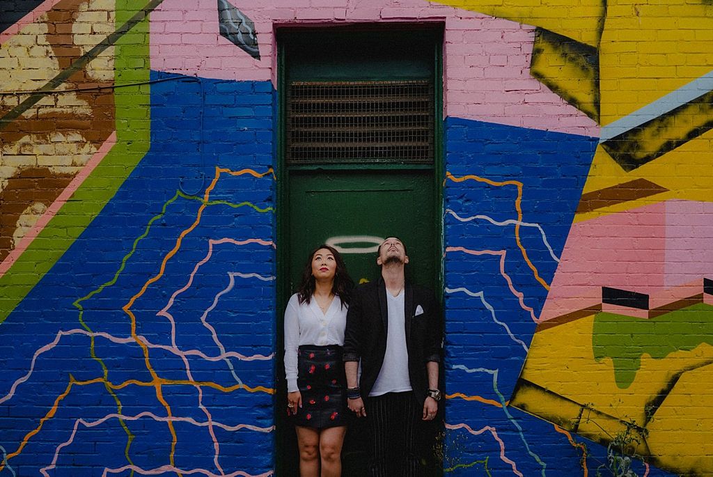 A couple poses in front of a colorful geometric street art mural with bold blue and yellow patterns.