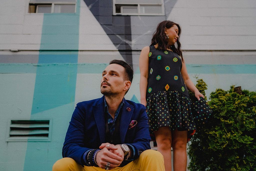 Stylish couple in contrasting outfits posing against modern architectural backdrop.