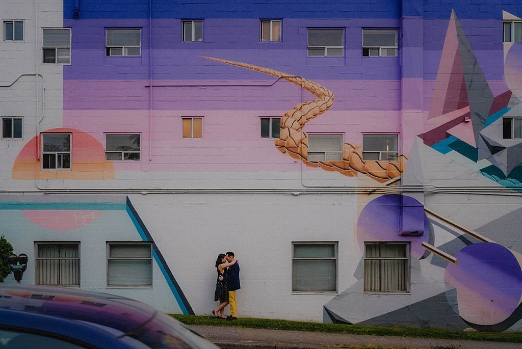 Couple embracing on staircase against pastel-colored building mural.