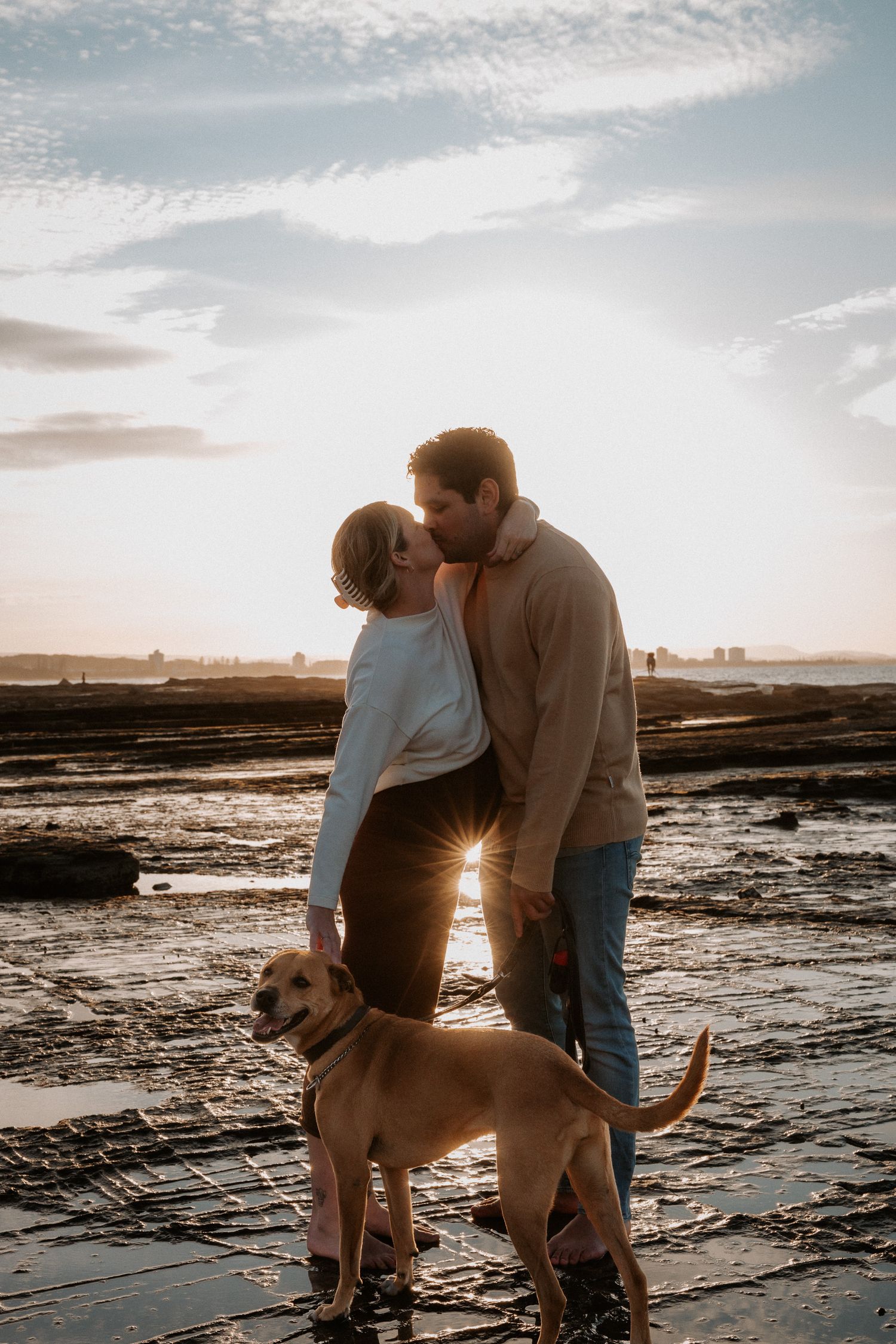 A couple shares an intimate moment at sunset on rocky beach terrain with their golden retriever.
