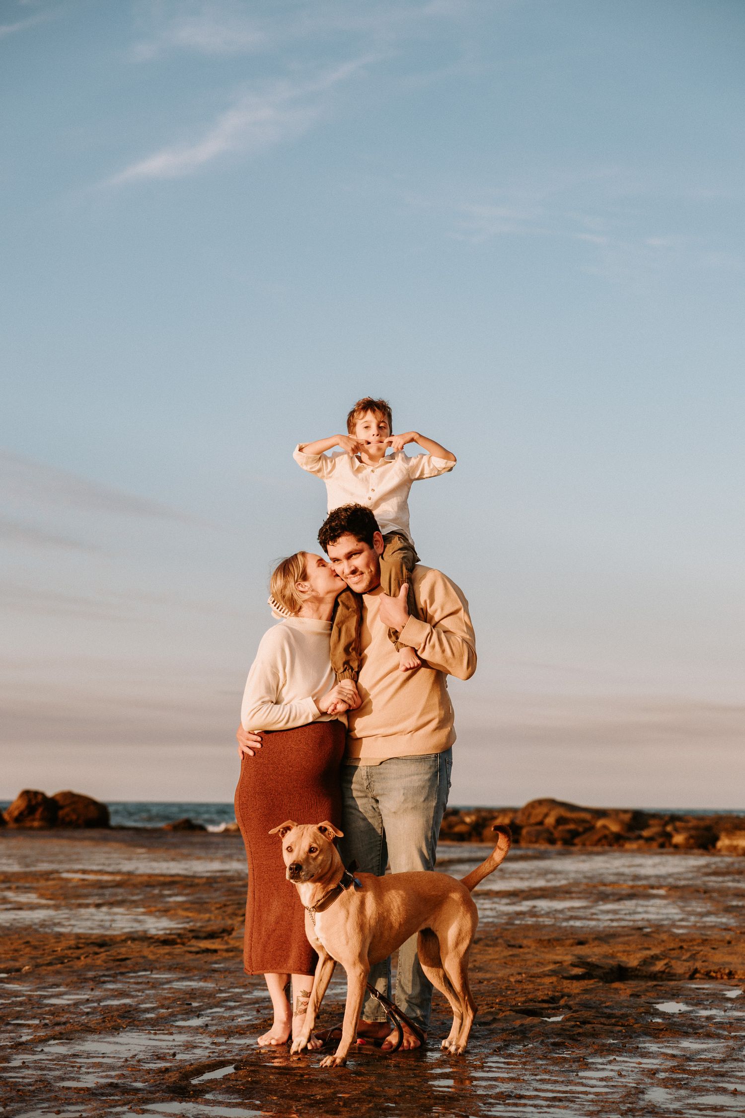 A sweet family moment captured on rocky beach terrain at sunset with their golden retriever.