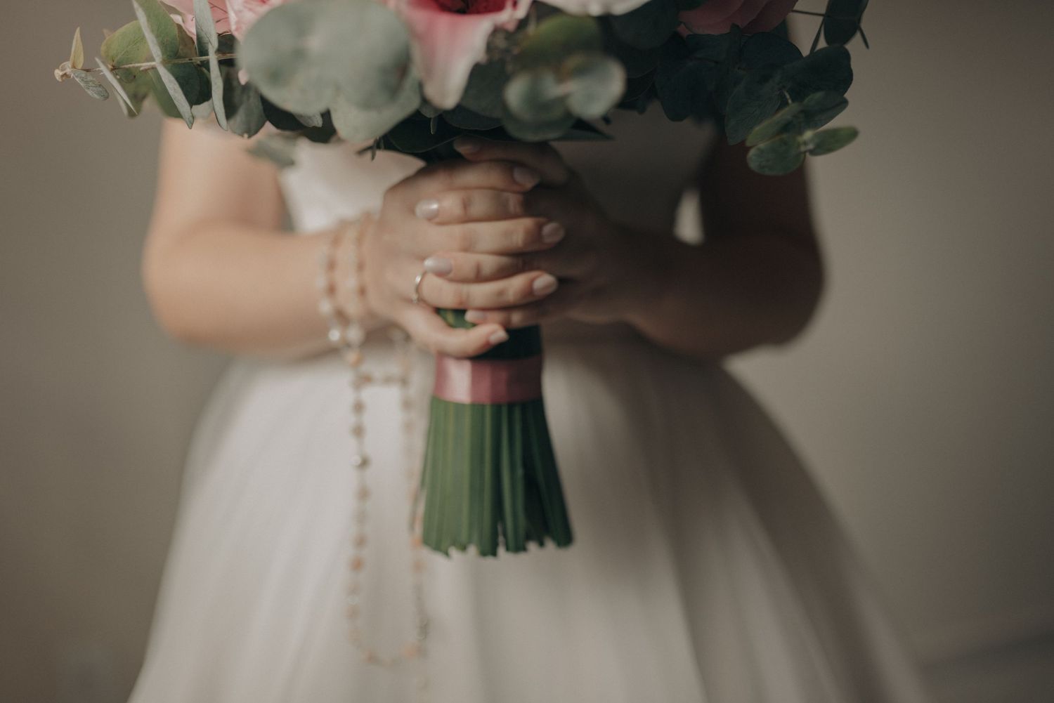 Close-up detail of hands holding a pink rose bouquet with pearl accents against a white wedding dress.