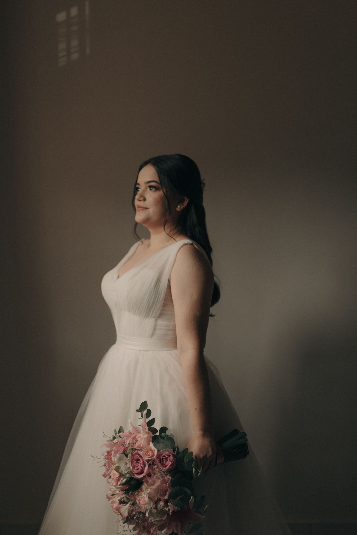 Elegant bridal portrait featuring a structured bodice gown and pink rose bouquet in moody lighting.