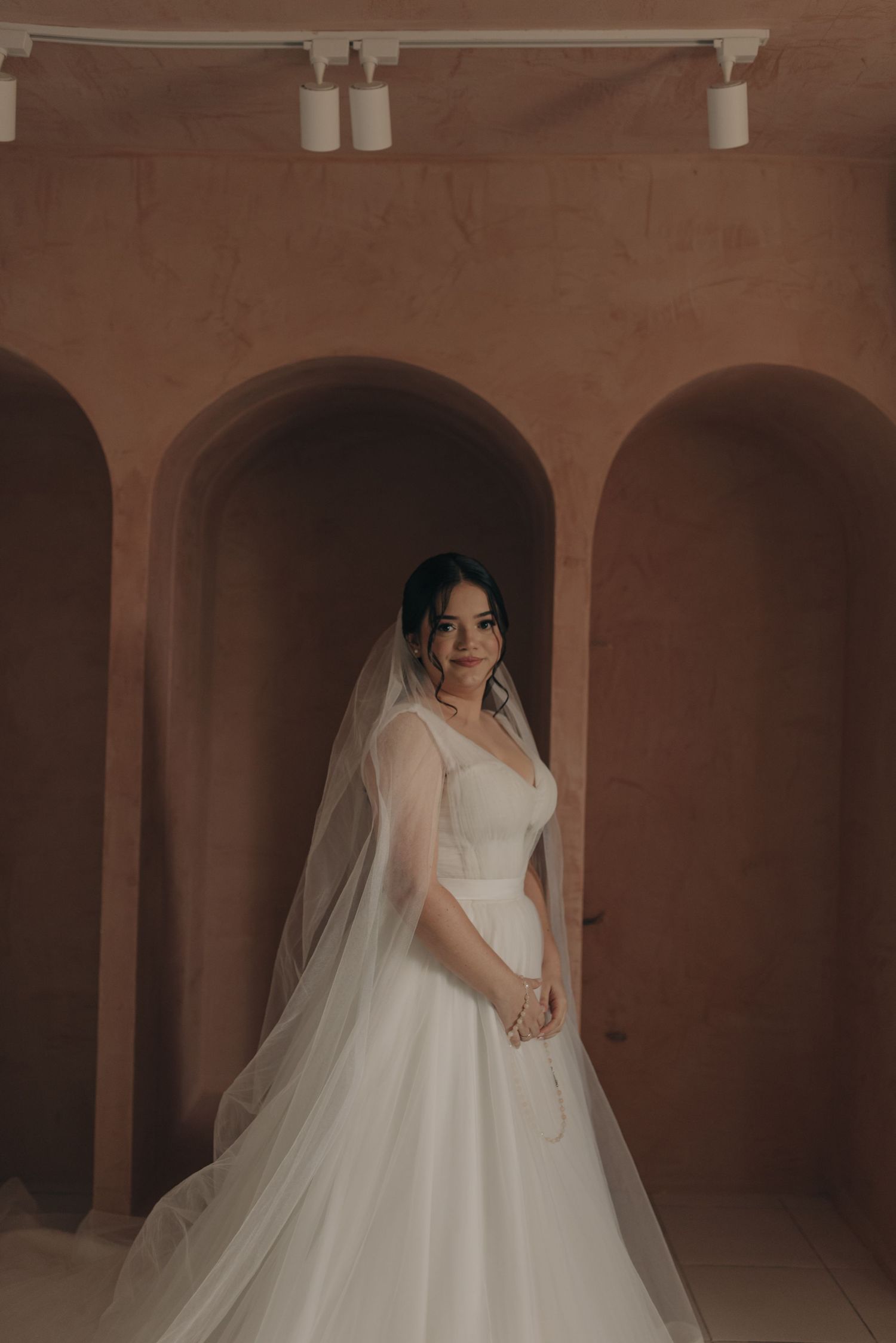 Bride in a flowing white gown and veil poses in an arched terracotta hallway.