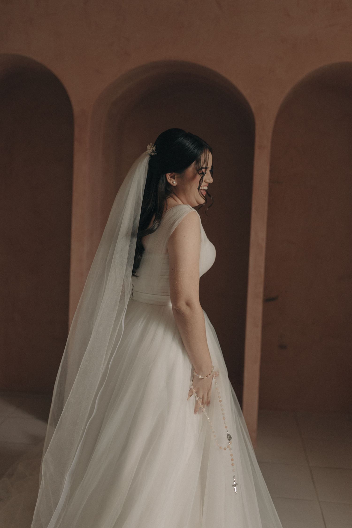 Side profile of a bride in a flowing wedding gown against terracotta archways.