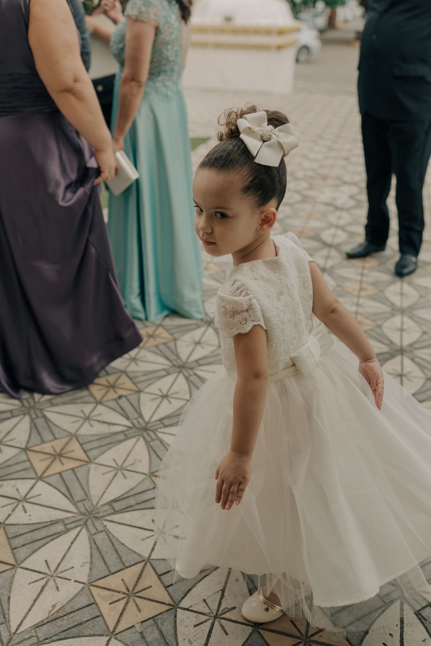 A young flower girl in a white dress with a bow hair accessory stands on a patterned tile floor at a formal event.