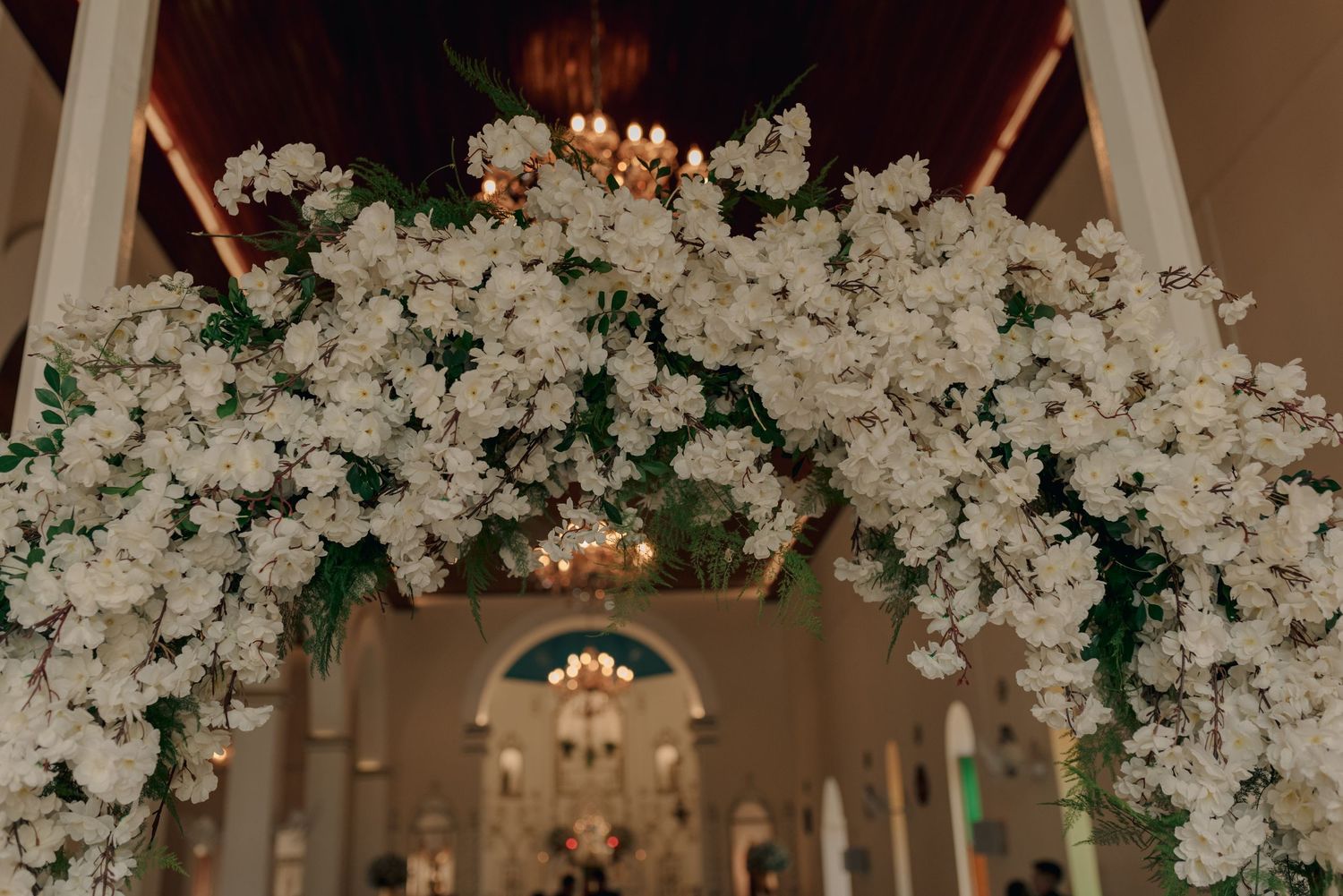 Elegant white floral arch with twinkling lights decorates the interior of a church for a wedding ceremony.